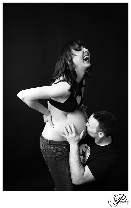 Lustiges Foto werdender Eltern, der Papa küsst den Babybauch und die Mama lacht herzlich. 